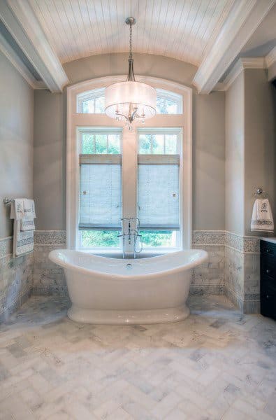 Freestanding bathtub beneath chandelier with large window and roman shades in luxury bathroom.