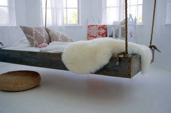 Cozy hanging bed with rustic wooden frame, white fur blanket, and soft cushions in a bright room.
