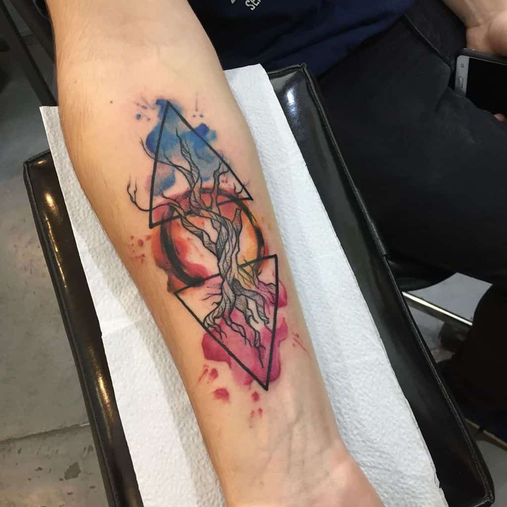 This tattoo blends geometric shapes with organic elements, featuring a tree within two overlapping triangles, enhanced by vibrant watercolor splashes. The design symbolizes balance, nature, and energy.