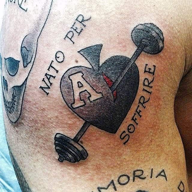 The tattoo features a barbell piercing through an ace of spades symbol, with the words "NATO PER SOFFRIRE" surrounding it