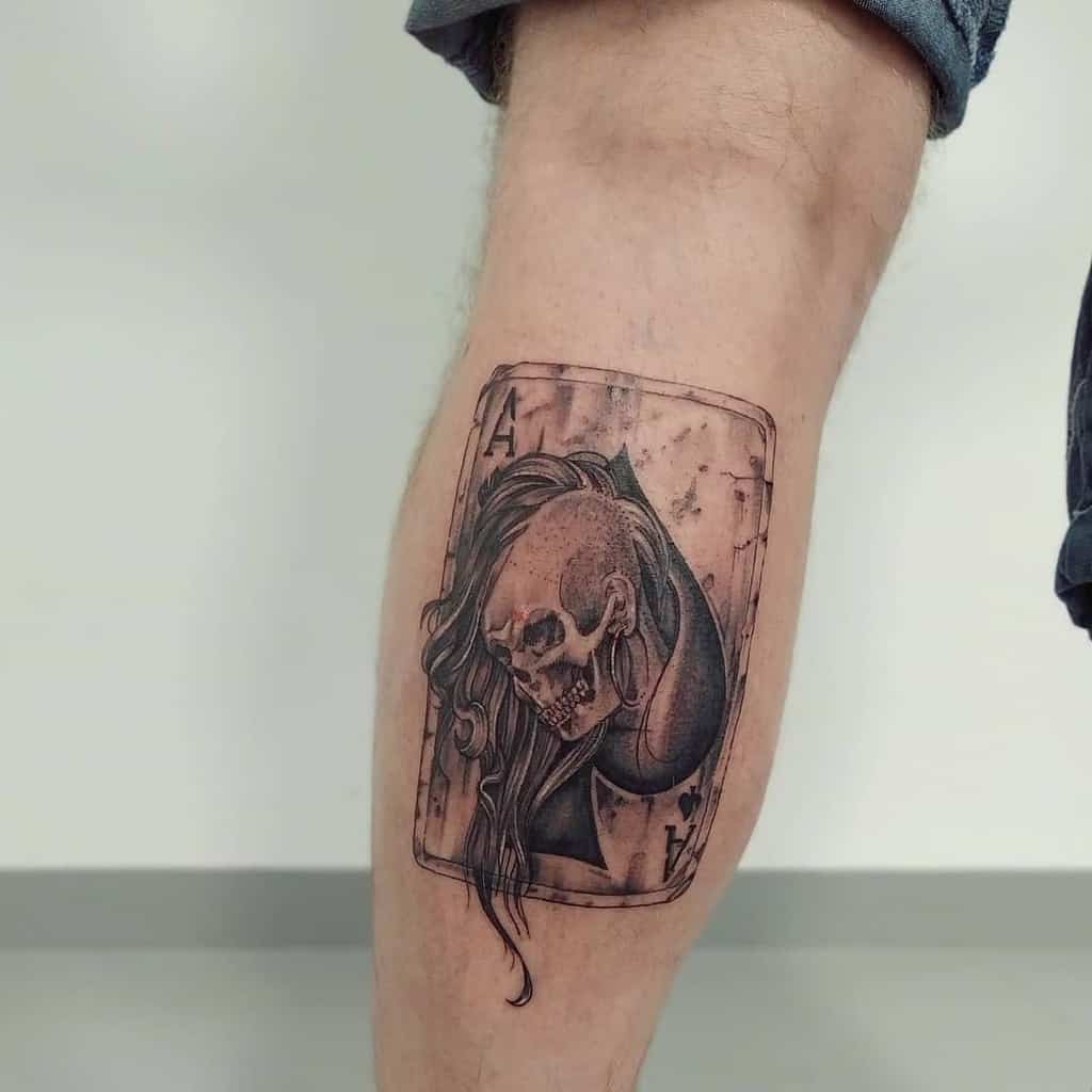 The tattoo features an ace of spades card with a skull wearing a large earring and long hair emerging from it