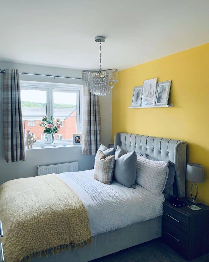 Cozy bedroom with a cheerful yellow accent wall, grey bed, chandelier, window view, and decorative pillows
