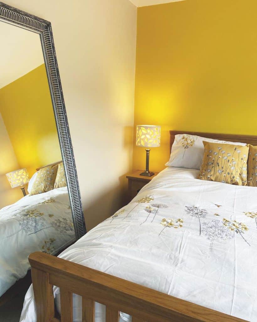 Cozy bedroom with a wooden bed, yellow floral bedding, a lamp on a nightstand, and a mirror reflecting the room