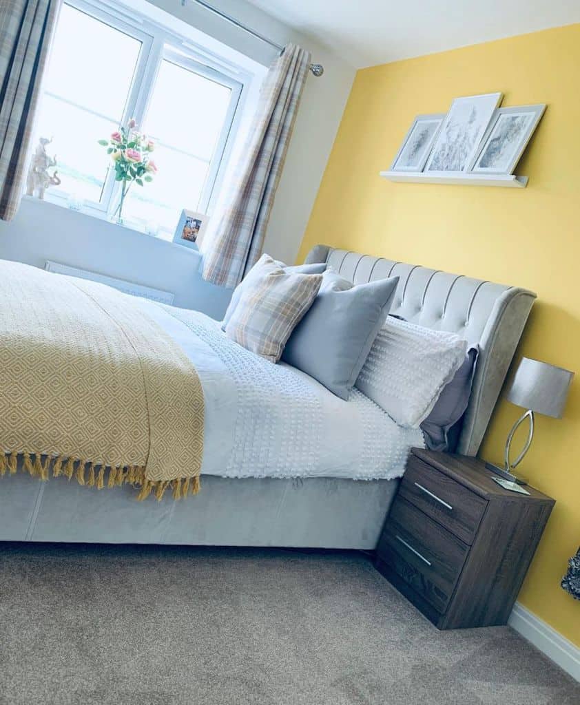 Cozy bedroom with yellow accent wall, bed with plush pillows and throw, bedside table, lamp, and a window view
