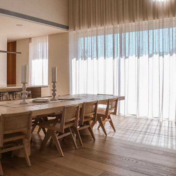Modern dining room with floor-to-ceiling sheer curtains, a rustic wooden table, and woven chairs.