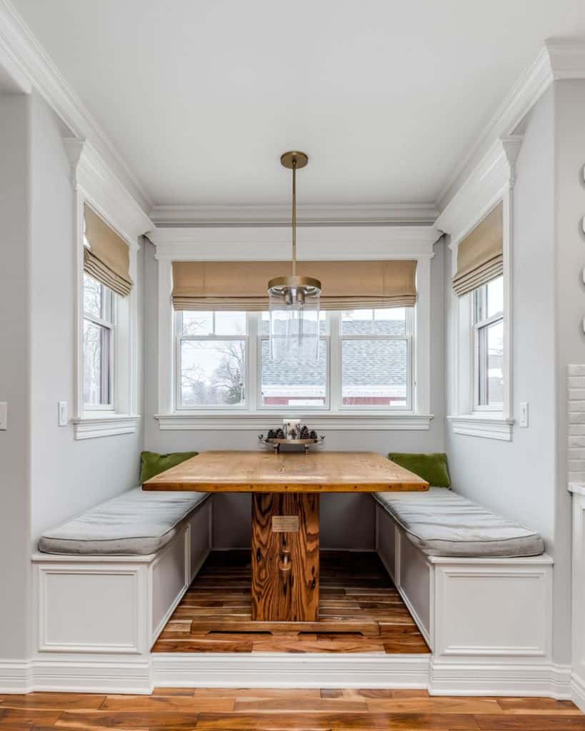 Cozy breakfast nook with wooden table, gray cushioned benches, and large windows.