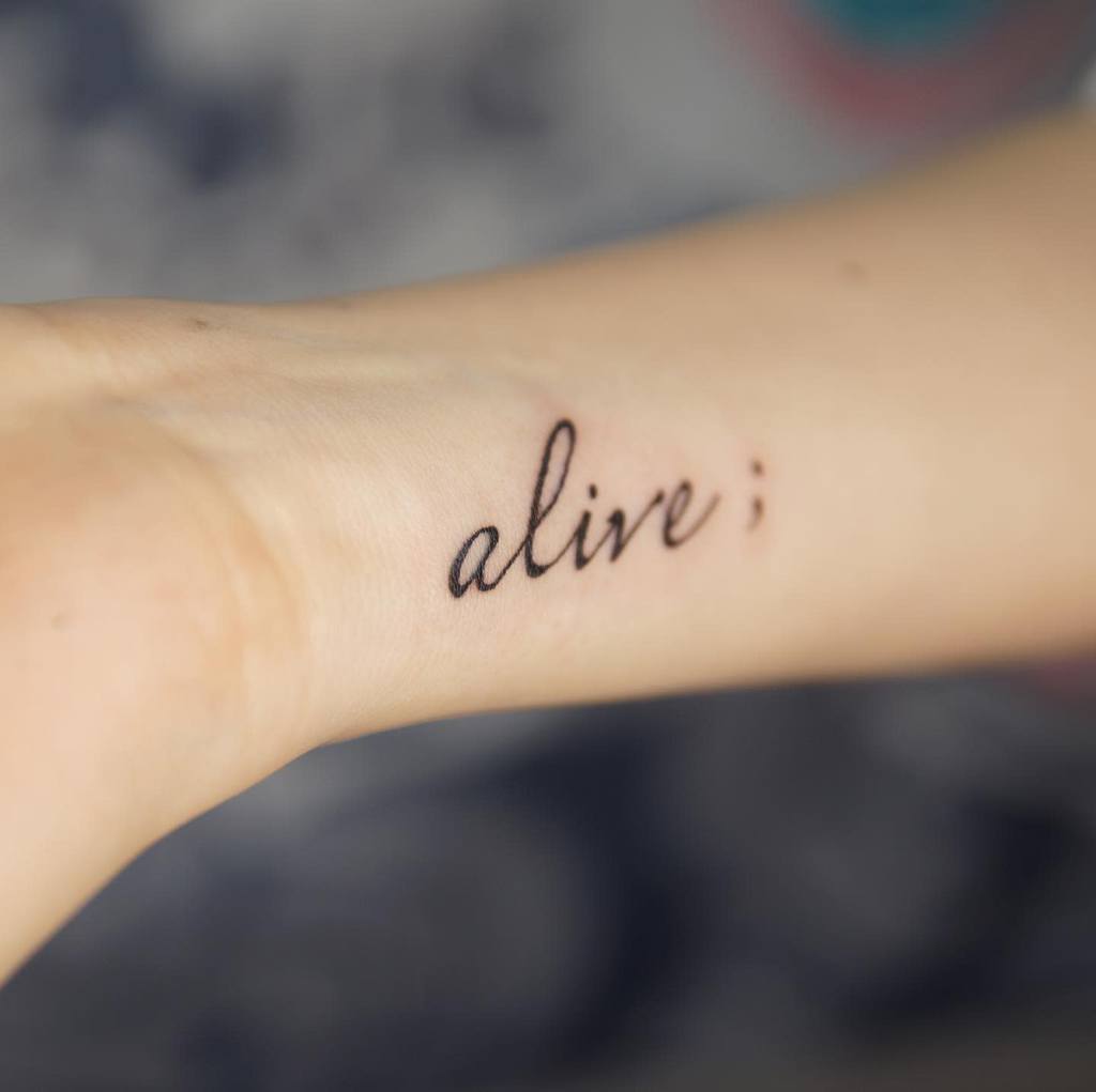 The tattoo features the word "alive" in elegant script with a semicolon at the end, symbolizing strength and resilience.