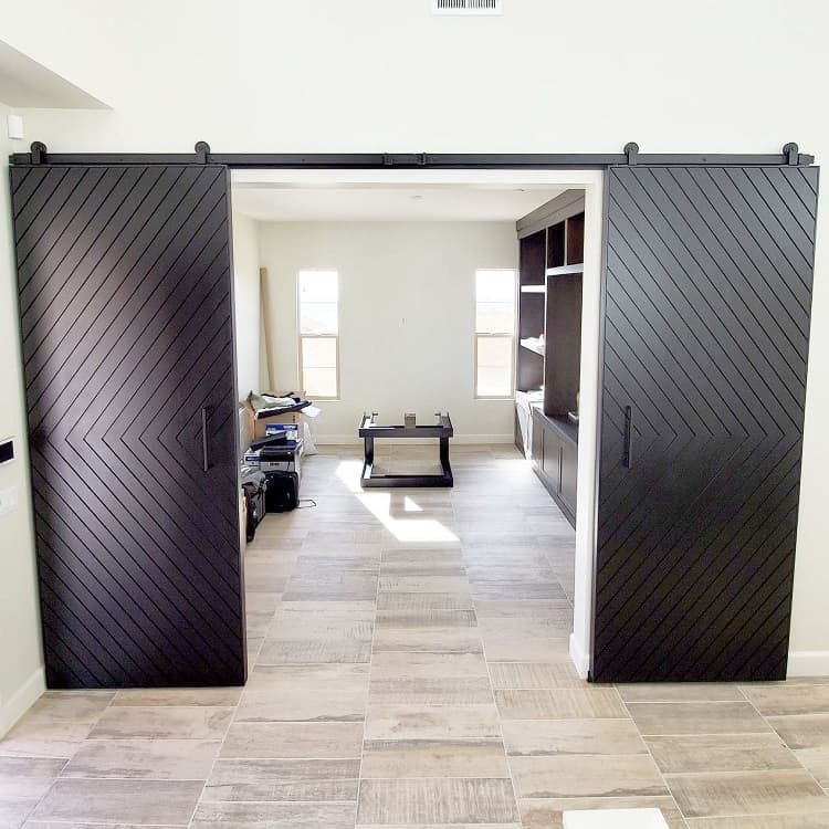 Black double sliding barn doors with chevron pattern in a modern room with tiled flooring and built-in shelving.