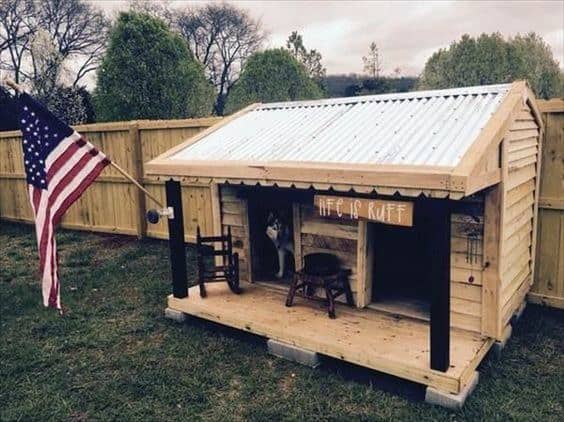 Wooden dog house with a metal roof, American flag, and a "Life is Ruff" sign, featuring two chairs and a dog inside