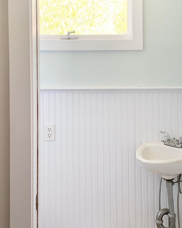 A small farmhouse bathroom corner featuring a white sink, mint green walls, wainscoting, a window, and an electrical outlet