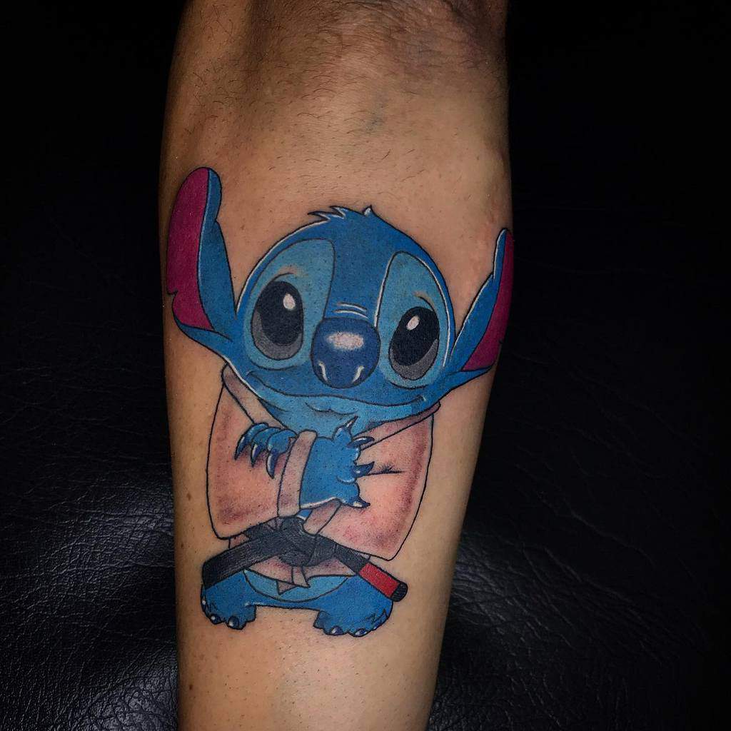 Colorful tattoo of Stitch from Lilo & Stitch wearing a Jedi robe and holding a lightsaber, with a playful expression, on the lower leg.