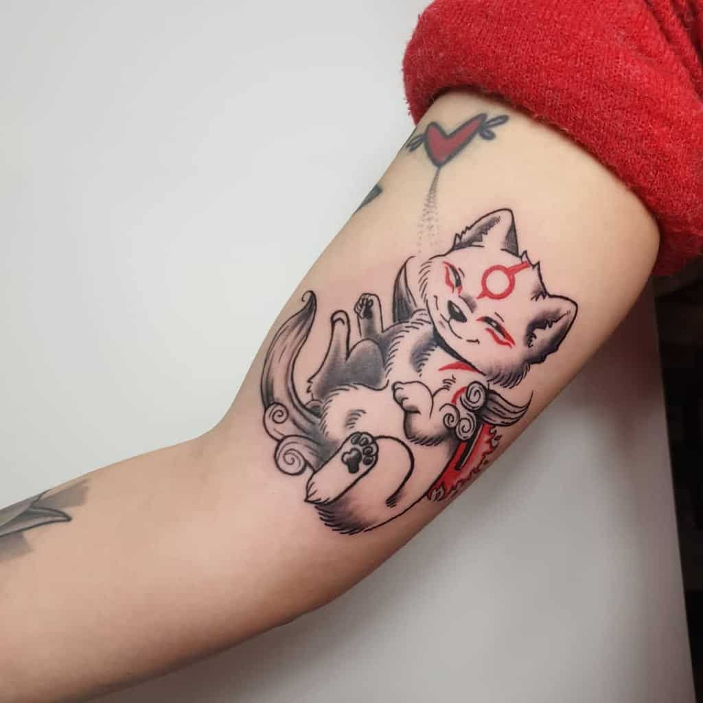 Adorable Okami tattoo of a chibi-style Amaterasu playfully curled up, featuring soft shading, red divine markings, and flowing mystical details