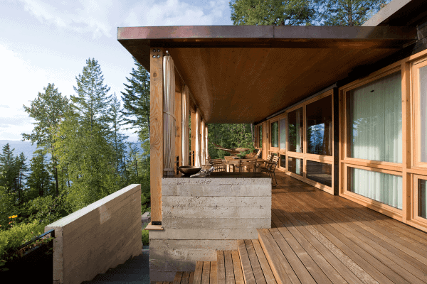 Modern wooden deck with large glass windows, outdoor seating, and forest views.