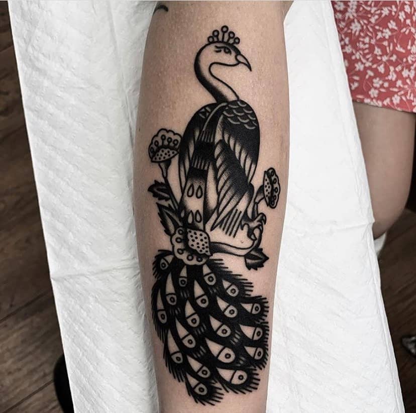 Black and white peacock tattoo with geometric feather details and eye patterns, accented by flowers, on the forearm.