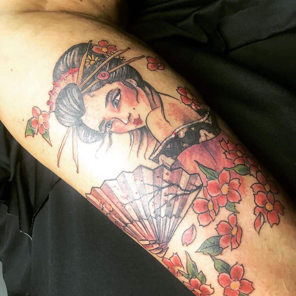 Colorful tattoo of a Geisha with intricate hair ornaments, holding a fan, surrounded by cherry blossoms in a traditional Japanese art style.