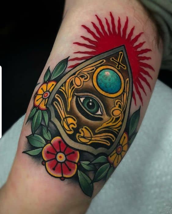 Tattoo of an ornate all-seeing eye inside a decorative planchette, surrounded by flowers and leaves, with a red radiating background, inked in vibrant colors on the forearm.
