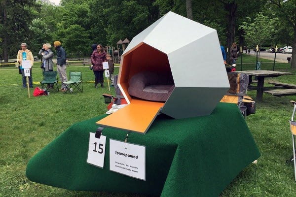 Geometric modern dog house with a sleek, angular design and an orange entrance, placed on a grassy area at an event