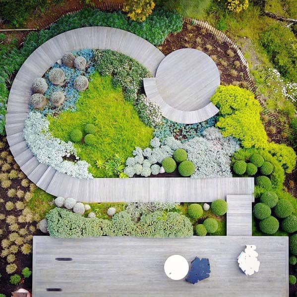 Aerial view of a garden with circular pathways, lush greenery, neatly trimmed shrubs, and a solitary bench