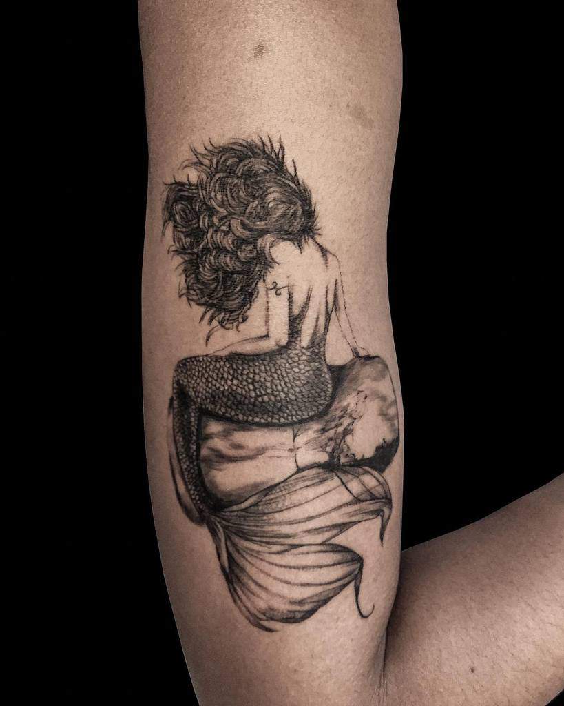 Black and grey tattoo of a mermaid with flowing hair sitting on a rock, gazing away, with intricate shading and fine details.