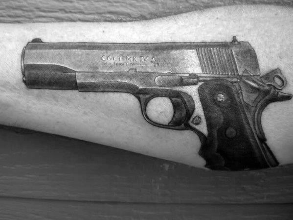 Realistic black and gray tattoo of a Colt MK IV 1911 pistol on the forearm, featuring fine details and shading for a three-dimensional effect.