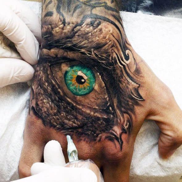 A realistic tattoo of a dragon's eye with a green iris is being inked on a person's hand by a tattoo artist wearing gloves