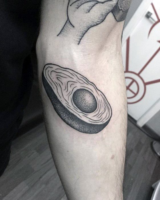 Black and grey avocado tattoo on inner arm with detailed shading and seed in center, designed in a bold, minimalist style.
