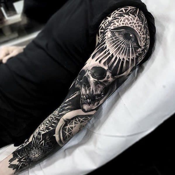 Black and grey full-sleeve tattoo featuring a skull, geometric patterns, and an all-seeing eye with radiating lines on the shoulder.