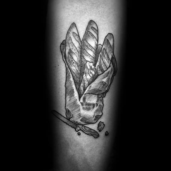 Detailed black and gray tattoo of baguettes wrapped in paper with a knife and crumbs below, creating a rustic and artistic bakery-inspired design.