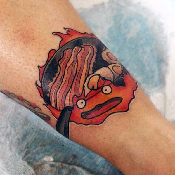 Calcifer tattoo design with bold outlines, featuring bacon and eggs in a frying pan above his fiery, expressive face.