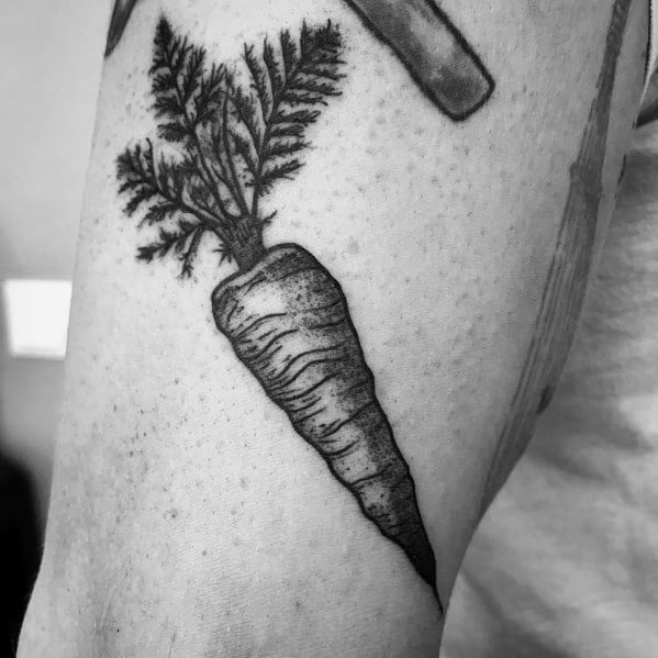 Realistic black and gray carrot tattoo on the upper arm, featuring detailed shading and leafy greens for a bold yet natural look.