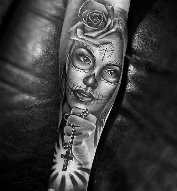 La Catrina tattoo featuring a woman with skull makeup, a rose on her head, and a hand holding a rosary with a cross.