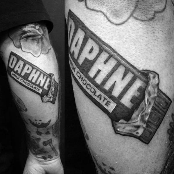 Black and gray tattoo of a personalized chocolate bar wrapper with the name Daphne, featuring a realistic design on the forearm.