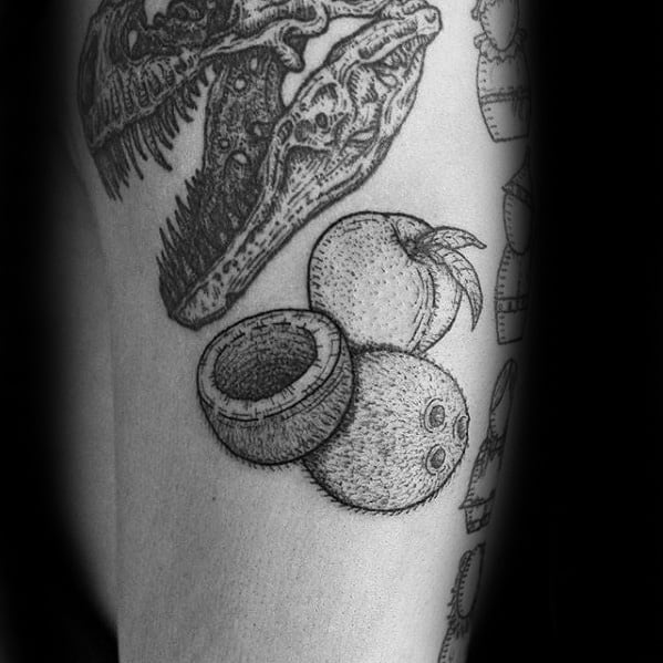 Detailed black ink coconut tattoo with a whole coconut, a halved one, and a dinosaur skull design above, placed on upper arm.
