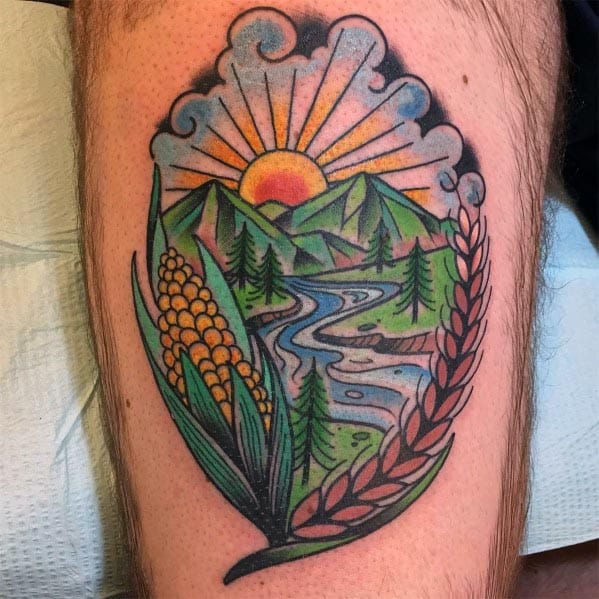 Colorful tattoo of a scenic landscape with mountains, a river, and a sunrise, framed by corn and wheat on a man's leg.