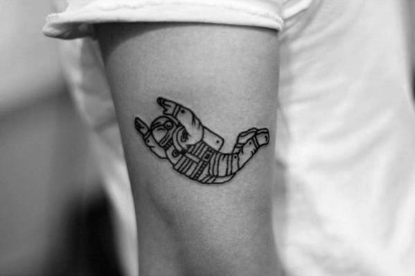 Minimalist tattoo of a falling astronaut with one hand doing the rock gesture, inked on the upper arm.