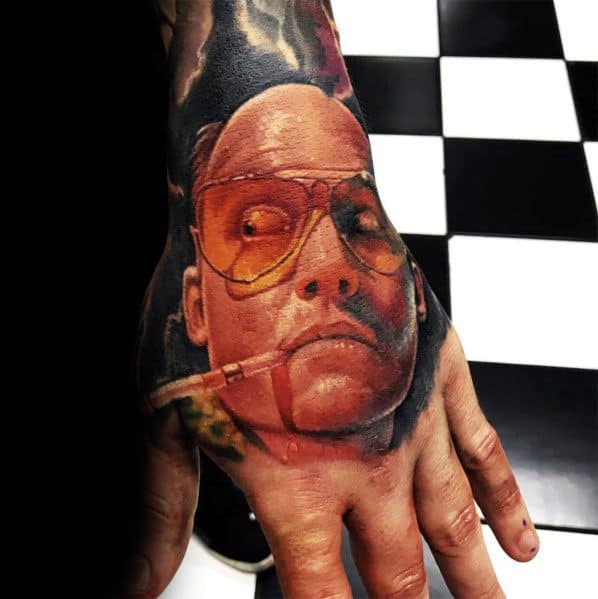 Hunter S. Thompson tattoo on a hand, featuring his face with orange-tinted aviator glasses and a cigarette, blending into a surreal background.