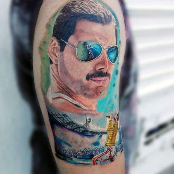Color tattoo of Freddie Mercury with reflective sunglasses, stadium scene below showing him in yellow jacket performing on stage.