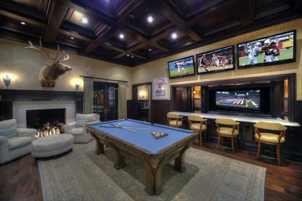 Luxurious game room with a pool table, cozy fireplace, and multiple TVs displaying sports, with bar seating