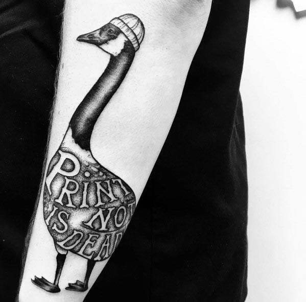 Black and white tattoo of a goose wearing a beanie, with the phrase "PRINT IS DEAD" written on its body, inked on a person's forearm.