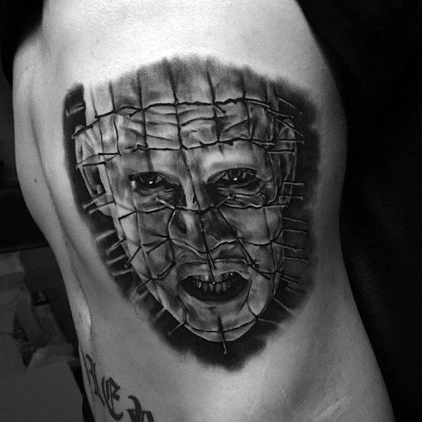Realistic Hellraiser-inspired tattoo of a terrifying face appearing behind a torn, stitched grid pattern, inked in black and grey.