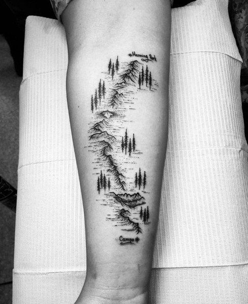 Hiking trail tattoo on forearm with detailed mountain range, trees, and labeled landmarks, symbolizing outdoor adventure.