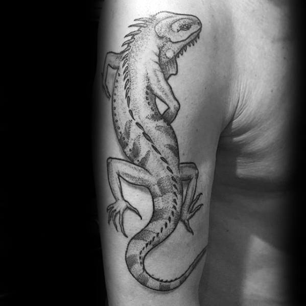 Iguana tattoo on upper arm featuring detailed black and grey shading with realistic scales and strong body posture.