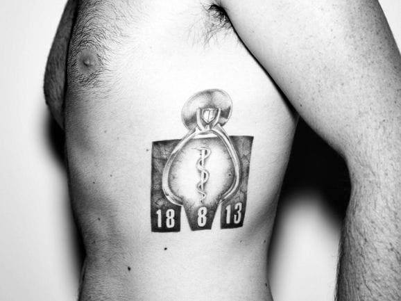Ironman logo tattoo with a medical symbol in the center, accompanied by the numbers "18 8 13," possibly marking a significant race or personal achievement.