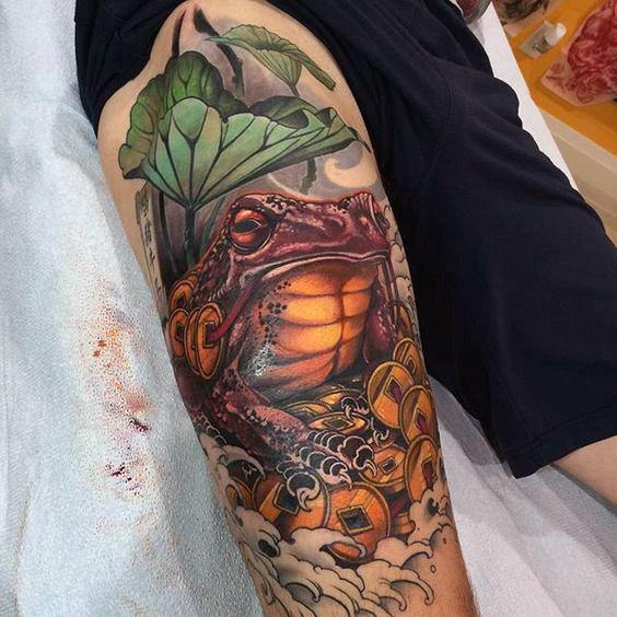 Japanese-style frog tattoo on a man's thigh, featuring a vibrant orange and red frog surrounded by coins, lotus leaves, and swirling clouds.