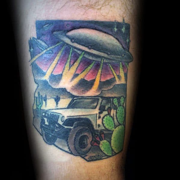 Colorful Jeep tattoo featuring an off-road Jeep in a desert landscape with cacti, a UFO beaming lights down, and a vibrant sunset sky.