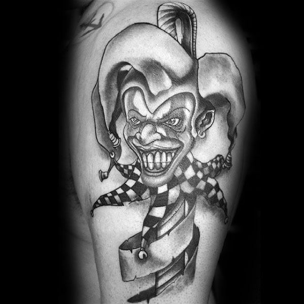 A black and gray tattoo of a smiling jester with a checkered outfit and exaggerated facial features, adorned with a flowing ribbon.
