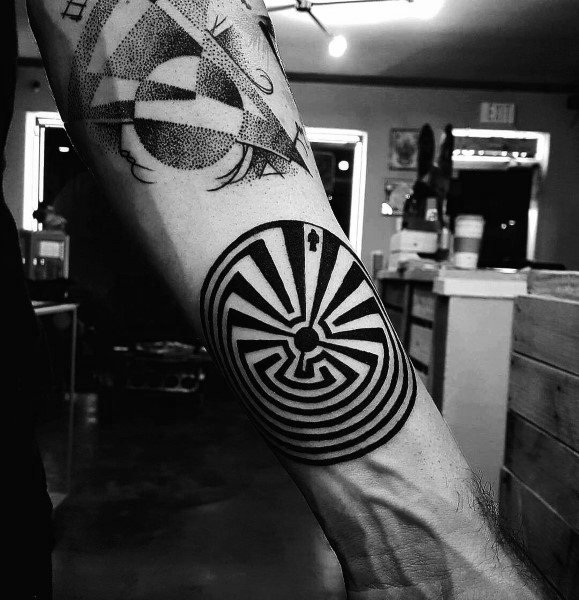 Black ink labyrinth tattoo on a man's forearm, featuring bold geometric lines forming a circular maze with a small figure at the entrance.