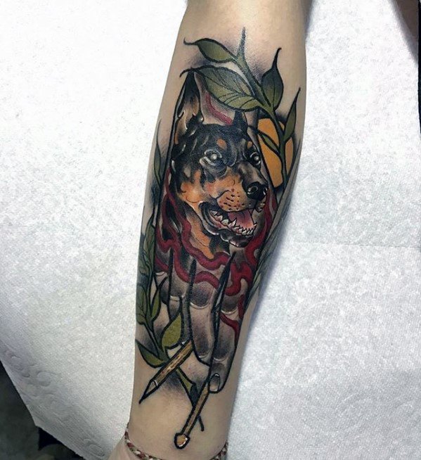 Doberman tattoo design featuring a realistic dog's head with fiery accents, surrounded by green leaves and geometric elements.
