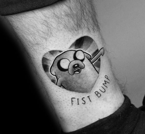 Black and grey Adventure Time tattoo of Jake the Dog in a heart shape with the phrase "Fist Bump" below, inked near the ankle.