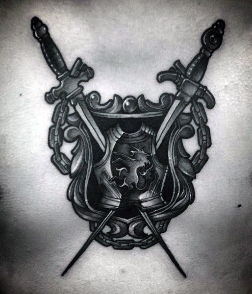 Black and grey tattoo of an ornate shield with a lion emblem, flanked by chains and crossed swords, creating a bold medieval design.